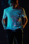 Modelo masculino andrógeno olhando em manchas brilhantes de luz vermelha e azul na camiseta, enquanto em pé sobre fundo preto — Fotografia de Stock