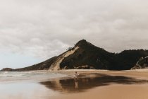 Человек ходит по мокрому песчаному берегу у морской воды на фоне гор и облачного неба — стоковое фото