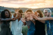 Группа разнообразных молодых друзей, улыбающихся и протягивающих руки к камере, стоя на размытом фоне удивительной сельской местности на закате — стоковое фото