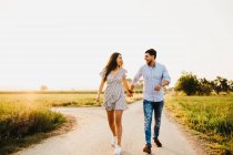 Joven y alegre hombre y mujer tomados de la mano y caminando en el campo a la luz del sol - foto de stock