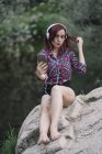 Red haired girl listens to music by the river — Stock Photo