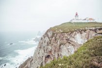 Пейзаж зеленой скалистой скалы с небольшим маяком над грубой водой океана в тумане, Португалия — стоковое фото