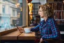 Vue latérale de la femme en T-shirt à carreaux assis dans un café avec une tasse de café sur la table et en utilisant un téléphone mobile — Photo de stock