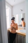 Jovem mulher topless em meias pretas e toalha na cabeça em pé no banheiro perto da janela e cobrindo o peito com os braços — Fotografia de Stock