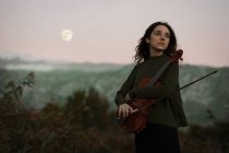 Adorável jovem senhora em vestido bonito segurando violino e olhando para longe enquanto estava em campo no fundo incrível de montanhas e céu — Fotografia de Stock
