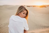 Молода жінка в білій футболці сидить на піску на заході сонця і дивиться через плече — стокове фото