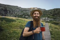 Молодий чоловік стоїть на шляху в горах з чашкою і дивиться на камеру — стокове фото