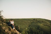 Nettes Paar, das sich umarmt, während es am felsigen Hang vor dem Hintergrund des wunderschönen Tales und der Berge sitzt — Stockfoto