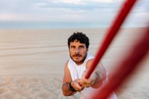 Uomo barbuto in abbigliamento sportivo tirando corda mentre si esercita sulla spiaggia di sabbia — Foto stock