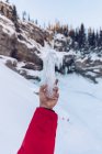 Cortar la mano en la chaqueta de invierno roja sosteniendo pedazo de hielo de cristal en el fondo de las montañas en la nieve, Canadá - foto de stock