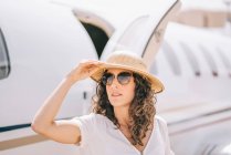 Jolie femme avec lunettes de soleil et chapeau à côté d'un avion. — Photo de stock