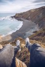 Geschnittenes Bild einer Person, die an einem bewölkten Tag in Asturien, Spanien, auf einer Klippe in der Nähe einer Bucht sitzt, mit wehendem Meerwasser — Stockfoto