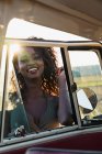Прекрасная афроамериканка улыбается и смотрит в камеру через окно винтажного автомобиля, проводя время на природе в солнечный день — стоковое фото