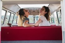 Vista trasera de dos mujeres riendo mientras se sientan en el asiento trasero de la furgoneta retro en la naturaleza - foto de stock