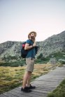 Joven barbudo de sombrero con mochila de pie en camino de madera que conduce a las montañas — Stock Photo