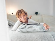 Очаровательный мальчик с вьющимися волосами, смотрящий в камеру, лежа на удобном диване рядом с подушками с цифровым планшетом — стоковое фото