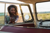 Bella donna afro-americana sorridente e guardando la fotocamera attraverso il finestrino di auto d'epoca mentre trascorreva del tempo nella natura nella giornata di sole — Foto stock