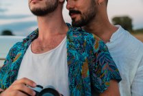 Beijar homens apaixonados abraçando ternamente enquanto estão de pé com câmera de fotos na natureza desfrutando de viagens — Fotografia de Stock