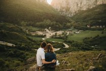 Милая пара обнимает и выносливые лбы, сидя на скалистом склоне на фоне красивой долины и гор — стоковое фото