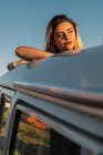 Привлекательная молодая женщина, опирающаяся на дверь машины и смотрящая в камеру, стоя в красивой природе в солнечный день — стоковое фото