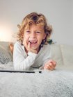 Fröhlicher Junge mit digitalem Tablet liegt zu Hause auf der Couch und lacht — Stockfoto