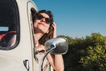 Счастливая молодая женщина в стильных солнцезащитных очках, выглядывающая из окна автомобиля, наслаждаясь летним солнцем на фоне зеленых деревьев и голубого неба — стоковое фото