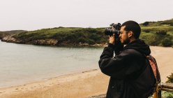 Side view of handsome guy with backpack standing on sandy coast and taking pictures of beautiful ocean during trip in nature — Stock Photo