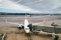 From above view of white airplane parked on landing ground with finger fixed to entrance on background of cloudy sky — Stock Photo