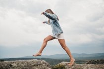 Side view of woman in dress and denim jacket leaping over crack jumping from rock on rock against cloudy sky — Stock Photo