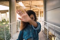 Jeune femme afro-américaine aux cheveux bouclés en denim debout dans un bâtiment de construction et se cachant face derrière la main — Photo de stock