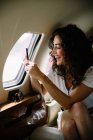 Jeune femme brune prenant des photos de vue extérieure à travers la fenêtre de l'avion et souriant joyeusement — Photo de stock