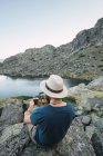 Young man sitting on rocks near lake with cup and using smartphone — Stock Photo