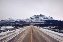 Ponte recintato con cartelli che attraversano fitti boschi invernali e fiume ghiacciato su sfondo montano innevato con cielo grigio nuvoloso — Foto stock