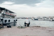 Вид сзади взрослого мужчины, сидящего на пирсе возле воды и кораблей и рыбалки в Стамбуле, Турция — стоковое фото