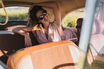 Bela mulher confiante em rosa e óculos de sol sentados no banco de trás no carro retro olhando para longe na luz solar — Fotografia de Stock