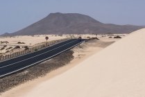 Larga carretera recta sobre llanura con dunas de arena con montañas, Islas Canarias - foto de stock