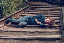 Одинокий очаровательный мальчик в джинсе спит на железной дороге, лежащей на деревянной балке в зеленой траве — стоковое фото