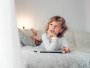 Вдумчивый мальчик лежит с цифровой табличкой на диване и смотрит в сторону — стоковое фото