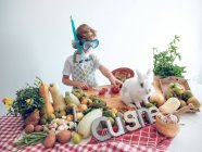 Jovem menino de pé e cozinhar legumes à mesa com coelho branco adorável — Fotografia de Stock