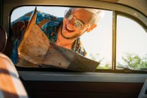 Красивый молодой человек с картой улыбаясь и глядя в винтажный автомобиль во время путешествия по сельской местности — стоковое фото