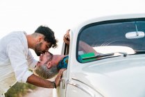 Vista lateral del hombre sentado dentro del coche y asomándose por la ventana besándose con su novio parado fuera en verano - foto de stock