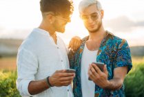 Two smiling men hugging and browsing smartphones while standing in beautiful countryside together — Stock Photo