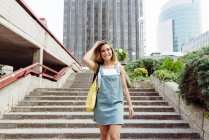 Sorridente giovane donna che regola i capelli e cammina al piano di sotto mentre guarda la fotocamera in città — Foto stock