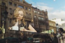 Жінка використовує смартфон у кафе за віконною панеллю з відображенням будівлі — стокове фото