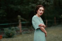 Side view of attractive young lady in elegant dress embracing herself and looking at camera while standing on blurred background of beautiful countryside — Stock Photo