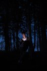 Красивий молодий хлопець спирається на стовбур дерева і переглядає смартфон, проводячи час у лісі вночі — стокове фото