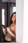 Joven mujer pensativa de pie cerca de la ventana y el uso de teléfono móvil - foto de stock