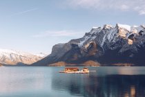 Edificio con tetto marrone sotto il sole in mezzo al lago altopiano con acqua blu chiaro su sfondo con pittoresche montagne e foresta — Foto stock