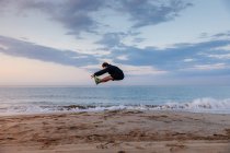 Homme actif en vêtements de sport sautant haut pendant l'entraînement en plein air sur la plage de sable au coucher du soleil — Photo de stock