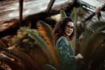 Vue latérale de belle jeune femme charmante afro-américaine en tissus verts et lunettes regardant la caméra et debout près des plantes — Photo de stock